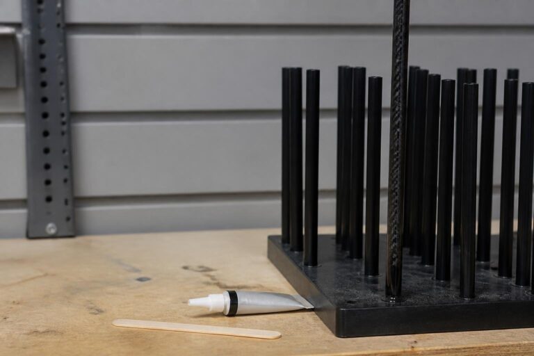 bright workshop lighting. A golf shaft standing upright in a drying rack with a small tube of epoxy and a mixing stick on the bench beside it
