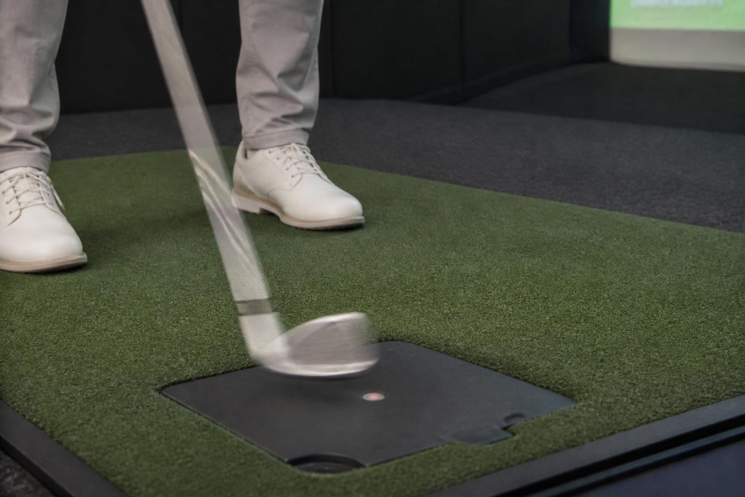 Ultra-realistic photograph of an iron clubhead being test-swung on a launch monitor mat in an indoor fitting bay, motion blur on the club